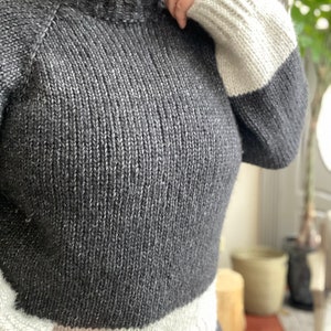 May include: A gray and white knit sweater with a mock neck and long sleeves. The sleeves have a white band at the cuff and the bottom of the sweater has a white band.