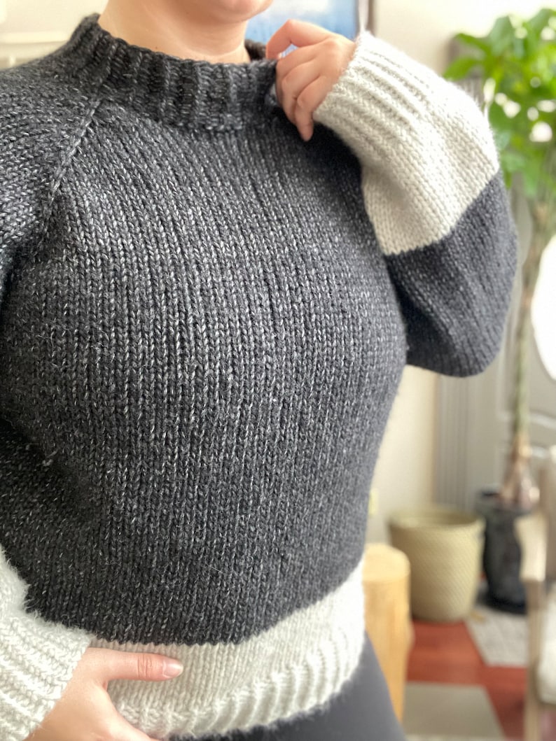 May include: A gray and white knit sweater with a mock neck and long sleeves. The sleeves have a white band at the cuff and the bottom of the sweater has a white band.