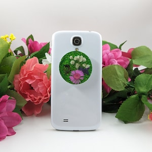 Peut inclure: Smartphone blanc avec un support circulaire vert pour téléphone orné de fleurs séchées. Le support a un fond scintillant et comprend des fleurs blanches, violettes et roses. Le téléphone est entouré de décorations florales roses et vertes.