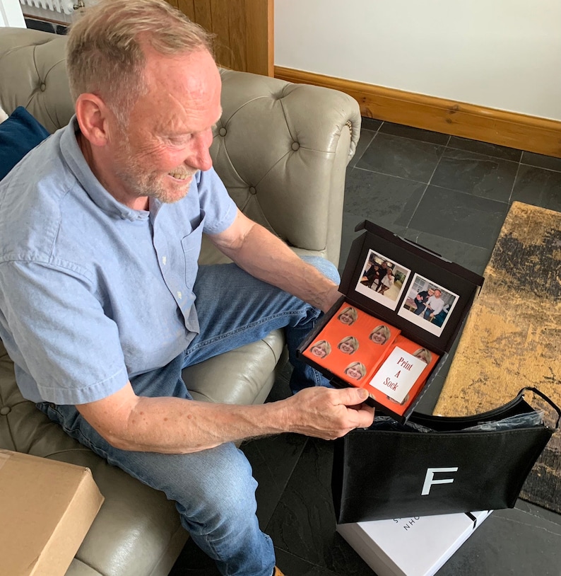K&ouml;nnte beinhalten: Eine Person &ouml;ffnet eine schwarze Box mit einem roten und wei&szlig;en gemusterten Papier darin. Die Box enth&auml;lt eine Reihe von Fotos und eine Karte mit der Aufschrift "Print A Stack".
