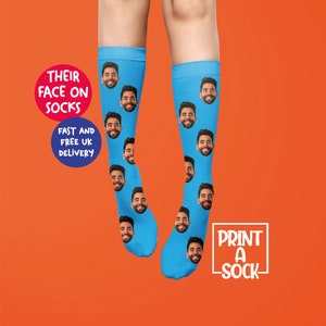 K&ouml;nnte beinhalten: Ein Paar blaue Socken mit einem sich wiederholenden Muster eines M&auml;nnergesichts. Der Text "DRUCKEN SIE EINE SOCKEN" befindet sich in einem wei&szlig;en Feld auf der rechten Seite des Bildes. Der Text "IHRE GESICHTER AUF SOCKEN" befindet sich in einem roten Kreis auf der linken Seite des Bildes. Der Text "SCHNELLER UND KOSTENLOSER VERSAND NACH GROSSBRITANNIEN" befindet sich in einem blauen Kreis unter dem roten Kreis.