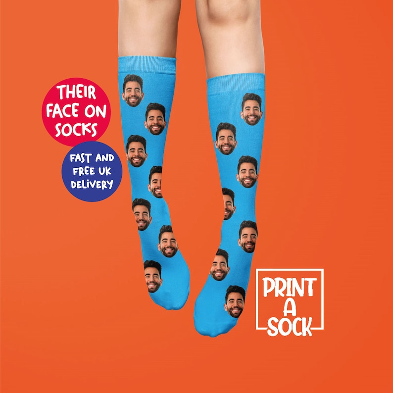 K&ouml;nnte beinhalten: Ein Paar blaue Socken mit einem sich wiederholenden Muster eines M&auml;nnergesichts. Der Text "DRUCKEN SIE EINE SOCKEN" befindet sich in einem wei&szlig;en Feld auf der rechten Seite des Bildes. Der Text "IHRE GESICHTER AUF SOCKEN" befindet sich in einem roten Kreis auf der linken Seite des Bildes. Der Text "SCHNELLER UND KOSTENLOSER VERSAND NACH GROSSBRITANNIEN" befindet sich in einem blauen Kreis unter dem roten Kreis.