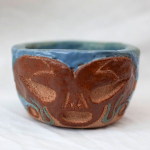 Pode incluir: Uma tigela pequena de cerâmica com um esmalte azul e um design marrom texturizado de um rosto. O rosto tem dois olhos, um nariz e uma boca.