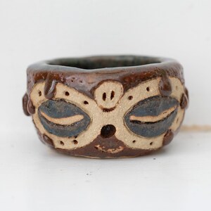 Op de afbeelding: Een kleine bruine keramische pot met een decoratief ontwerp van ogen en een mond. De pot heeft een witte glazuur en bruine accenten.