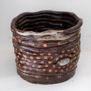 Könnte beinhalten: Ein brauner Keramiktopf mit einem gewellten Rand und kupferfarbenen Flecken. Der Topf hat eine hellgraue Glasur und einen einzigen ovalen Akzent in Hellblau.