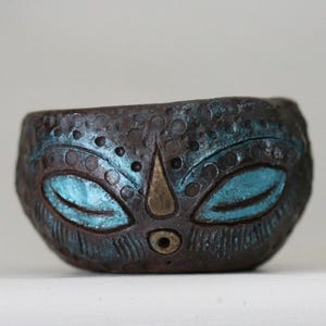 Puede incluir: Una pequeña maceta de cerámica marrón con un diseño de cara. La cara tiene ojos azules, una nariz marrón y un pequeño agujero de drenaje. La maceta está decorada con un patrón de pequeños puntos.