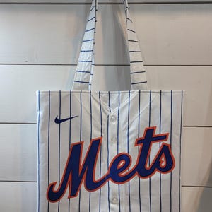 May include: White tote bag with blue and orange "Mets" lettering and a Nike logo. The bag has a striped design that resembles a baseball jersey.