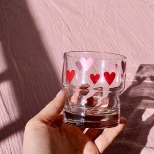 May include: A clear glass tumbler decorated with red and pink heart shapes. The glass has a rounded base and a slightly flared top. The hearts are evenly spaced around the glass, creating a festive and romantic design.