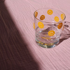 May include: A clear glass decorated with yellow and brown smiley faces. The glass contains a clear liquid and has the text "Good Day" in yellow. The glass is on a pink surface.