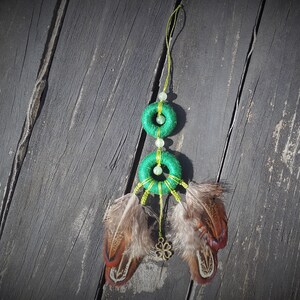 Peut inclure: Un attrape-rêves vert et or avec deux cercles et des plumes brunes qui pendent du bas.