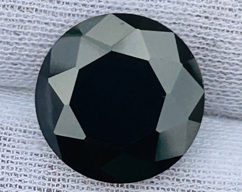 May include: A black round gemstone with a faceted cut. The gemstone is set on a white fabric background.