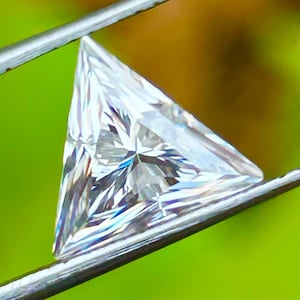 May include: A clear triangular diamond gemstone held by tweezers against a green background.