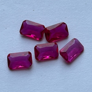 Puede incluir: Cinco gemas rectangulares de color rojo intenso con bordes facetados. Las gemas están dispuestas sobre una superficie de color claro, mostrando su color y claridad vibrantes. Estas gemas podrían usarse para la fabricación de joyas u otros fines decorativos.