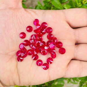 May include: A collection of loose, round, red gemstones. The gemstones are scattered on a hand.