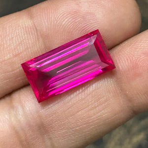 May include: A vivid, rectangular-cut, hot pink gemstone. The gem has a faceted surface that reflects light, creating a brilliant display. The stone is held in a hand, showcasing its colour and clarity. This gemstone could be used in jewellery.