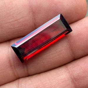 May include: A rectangular, faceted gemstone with a deep red hue. The gem has a clean, sharp cut with a polished surface, reflecting light. The gemstone is held in a hand, showcasing its color and clarity.
