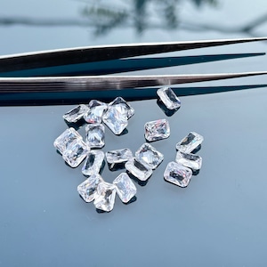 Könnte beinhalten: Eine Ansammlung klarer, rechteckig geschliffener Edelsteine, die auf einer reflektierenden Oberfläche verstreut sind. Die Edelsteine haben unterschiedliche Größen und Formen und weisen ein facettiertes Aussehen auf. Eine silberne Pinzette befindet sich darüber.