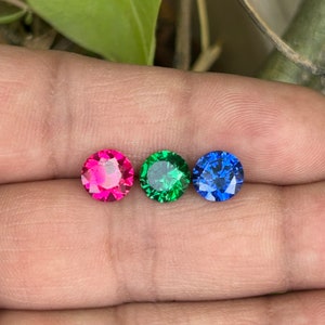 Peut inclure: Trois pierres précieuses rondes, une rose, une verte et une bleue, sont présentées sur un doigt.