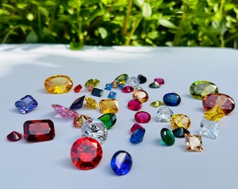 May include: Assortment of colorful gemstones in various shapes and sizes, including round, square, and oval. The gemstones are arranged on a white surface and include red, blue, green, yellow, and clear stones.