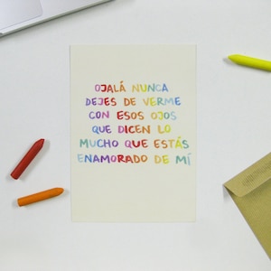 May include: A white card with a rainbow-colored message that reads "OJALÁ NUNCA DEJES DE VERME CON ESOS OJOS QUE DICEN LO MUCHO QUE ESTÁS ENAMORADO DE MÍ".