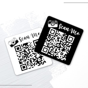 May include: Two square QR code stickers, one black and one white, with the text "Scan Me" in cursive and a camera icon. The QR codes are black and white, and the stickers are on a white background.