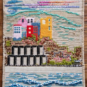 Può includere: Opera d'arte tessile cucita a mano raffigurante una scena di città costiera. L'opera presenta edifici colorati in rosa, rosso, blu e giallo, sullo sfondo di un cielo strutturato con tonalità blu, viola e rosa. La parte inferiore mostra un oceano stilizzato.