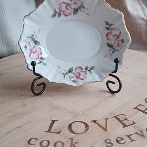 May include: A white porcelain saucer with a gold rim and pink floral design. The saucer is on a black metal stand and a wooden surface with the text "LOVE cook. serve."