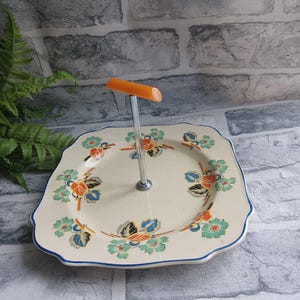 May include: A vintage cake stand with a square, scalloped plate and a metal stand with an orange plastic handle. The plate has a floral design in shades of blue, green, orange, and yellow.