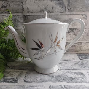 May include: White porcelain teapot with a silver and brown bamboo design. The teapot has a lid and a handle.
