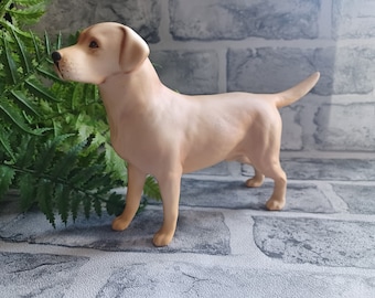 May include: A light tan Labrador Retriever figurine. The dog is standing, facing left, with its tail slightly raised. The figurine has a smooth finish and is set against a brick-like background with green foliage.