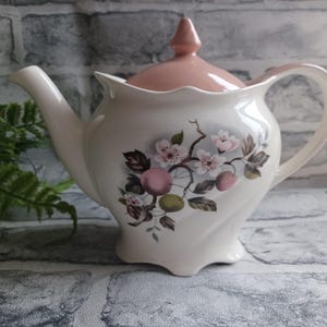 May include: A white teapot with a pink lid and a floral design featuring pink flowers and green leaves. The teapot has a curved handle and a spout.