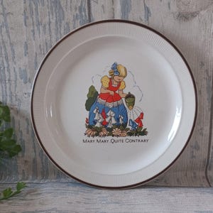 May include: White ceramic plate with a brown rim and the text "MARY, MARY, QUITE CONTRARY". The plate features a colorful illustration of a girl watering flowers. The girl is wearing a red dress and blue skirt.