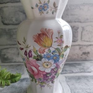 Może przedstawiać: Biały ceramiczny wazon z kwiatowym wzorem. Wazon ma falisty brzeg i złotą obwódkę wokół podstawy. Kwiaty są różowe, żółte, niebieskie i fioletowe.