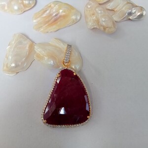 Può includere: Un ciondolo con una pietra preziosa di colore rosso intenso, con una montatura in oro e un bordo di piccole pietre trasparenti.