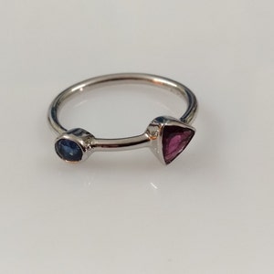 May include: A silver ring with a small blue gemstone on one side and a triangular pink gemstone on the other side.
