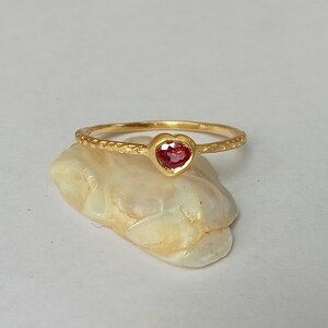 Puede incluir: Un delicado anillo de oro con una banda texturizada y una pequeña piedra preciosa rosa en forma de corazón.