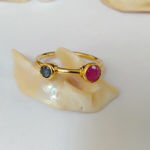 Op de afbeelding: Een gouden ring met een donkerblauwe edelsteen en een roze edelsteen. De ring wordt getoond op een witte schelp.