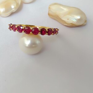 May include: A gold ring set with vibrant red gemstones. The ring is displayed on a white surface, surrounded by several large, irregularly shaped pearls. The gemstones are evenly spaced around the ring's band.