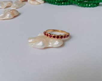 May include: A gold ring with a band of red gemstones. The ring is resting on a white, pearlescent surface.