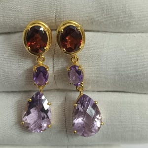 May include: Gold dangle earrings with three gemstones. The top gemstone is a round, dark red garnet. The middle gemstone is a round, light purple amethyst. The bottom gemstone is a pear-shaped, light purple amethyst.