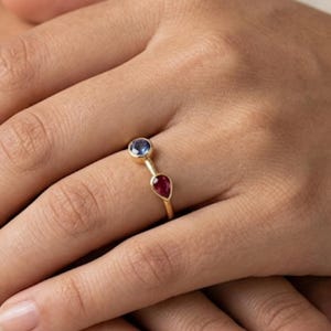 May include: A gold ring with a round blue gemstone and a pear-shaped red gemstone. The ring is on a hand with manicured nails. The ring's design is simple and elegant, suitable for everyday wear.
