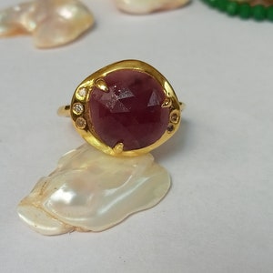 Puede incluir: Un anillo de oro con una gran piedra preciosa roja oscura facetada rodeada de piedras preciosas más pequeñas y transparentes.
