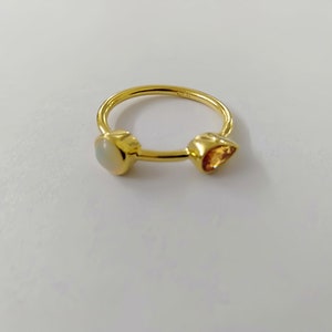May include: A gold ring with two gemstones. One gemstone is oval-shaped and white, while the other is a triangular, orange-yellow color. The ring is set against a white background, highlighting the jewelry's design and materials.