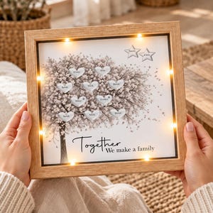 Puede incluir: Un marco de madera cuadrado que contiene una pantalla iluminada con una ilustración de un árbol con hojas en forma de corazón con nombres, dos estrellas en la parte superior y el texto "Together We make a family" en fuente negra.