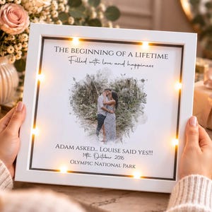 Op de afbeelding: Een vierkante witte LED-lichtbak met een centraal hartvormige foto van een koppel. De tekst luidt: THE BEGINNING OF A LIFETIME, Filled with love and happiness, ADAM ASKED... LOUISE SAID YES!!!, 14th October 2016, OLYMPIC NATIONAL PARK.