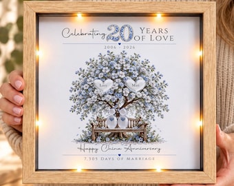 Puede incluir: Un marco cuadrado de madera con luces LED integradas que contiene una impresión de un árbol con flores azules, dos piedras en forma de corazón con los nombres Lucy y Matt, y dos figuras de piedra en un banco. El texto dice Celebrating 20 Years of Love.