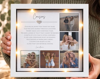 May include: A square white shadow box frame with integrated LED lights, featuring a collage of four photographs and a printed poem titled Cousins. The text includes lines about a special bond, shared laughter, and lifelong friendship.