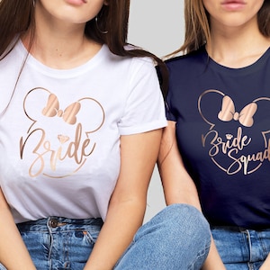 May include: Two white and navy blue t-shirts with a rose gold Mickey Mouse head design. The white shirt says "Bride" and the navy shirt says "Bride Squad".