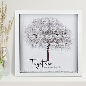 Op de afbeelding: Een witte ingelijste schaduwdoos met een zilveren glitterachtergrond en een witte boom met zilveren harten. Elk hart heeft een naam erop geschreven. De tekst "Together is our favourite place to be." staat onder de boom.