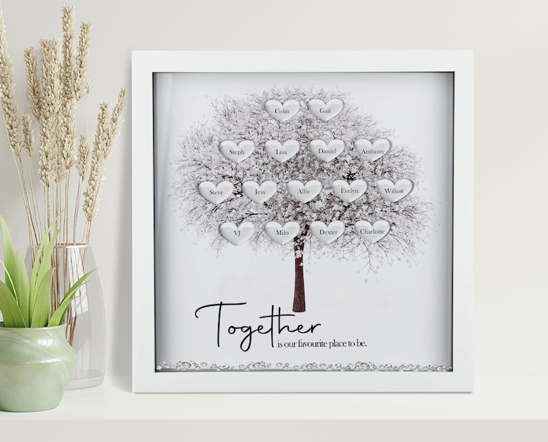 Op de afbeelding: Een witte ingelijste schaduwdoos met een zilveren glitterachtergrond en een witte boom met zilveren harten. Elk hart heeft een naam erop geschreven. De tekst "Together is our favourite place to be." staat onder de boom.
